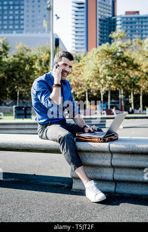 Smiling businessman on the phone sitting on bench outdoors with laptop Stock Photo