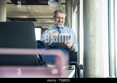 Smiling mature businessman sitting on chair using tablet Stock Photo
