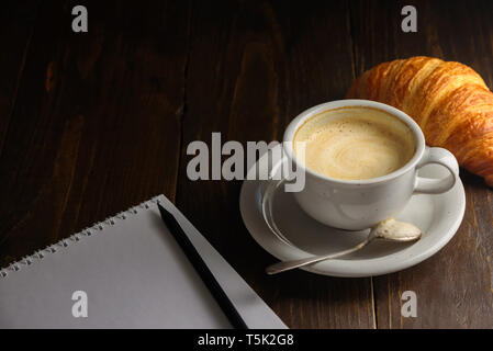 Cappuccino with croissant on dark wooden background. Text space. Blank notepad and pencil near it Stock Photo