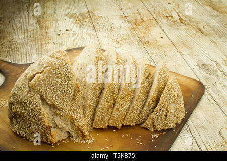 Close-up of a sliced  loaf of homemade bread with sesame seeds on wooden cutting board in selective focus on wooden table background Stock Photo