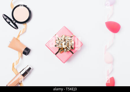 Pink gift box with golden bow among decorative cosmetics for even face skin tone and makeup sponges for its application, top view on white background. Stock Photo