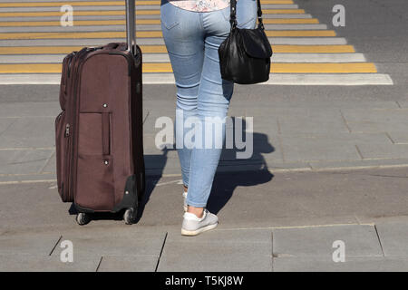 Woman with a suitcase on wheels stands on a pedestrian crossing, back view. Female legs in blue jeans on a crosswalk, concept of travel, waiting taxi Stock Photo
