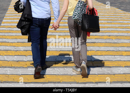 Couple in love walks on pedestrian crossing holding hands. Road zebra marking, people on a crosswalk in warm sunny weather, street safety concept Stock Photo