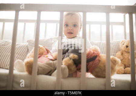 The toddler sits in a crib and plays among pillows and teddy bears. Stock Photo