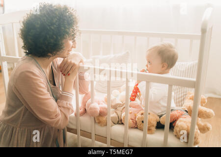 Happy mother sits next her baby with toys in the crib. Stock Photo