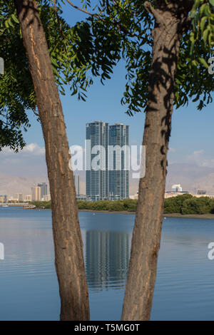 Looking past a pair of trees towards Julphar Towers, downtown, and Jebal Jais in Ras al Khaimah, United Arab Emirates. Stock Photo