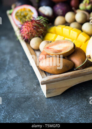 Various fresh Thai fruits - rambutan, mango, mangosteen, longan, sapodilla, passion fruit, salak, on a wooden tray and a gray background. Stock Photo