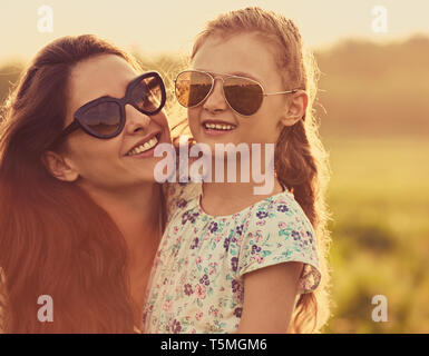 Happy fashion kid girl whisper the secret to her mother  in ear in trendy sunglasses in profile view on nature sunset background. Closeup toned portra Stock Photo
