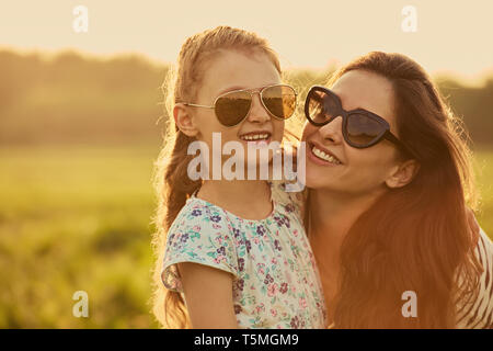 Happy fashion kid girl whisper the secret to her mother  in ear in trendy sunglasses in profile view on nature sunset background. Closeup Stock Photo