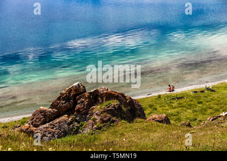A couple of young people with bicycles are resting on the shore of a mountain lake. Turquoise water reflects the sky with clouds. Idyll in nature. Mou Stock Photo