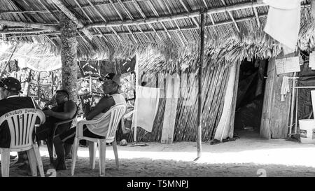 Guna (aka Kuna) tribal members relax under a large communal hut made of palm fronds, watching as tourists visit their island and shop for Molas Stock Photo