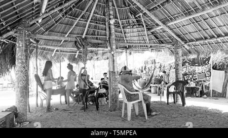 Guna (Kuna) tribal members relax under a large communal hut made of palm fronds, watching as tourists visit their island and shop for Molas Stock Photo