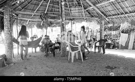 Guna (Kuna) tribal members relax under a large communal hut made of palm fronds, watching as tourists visit their island and shop for Molas Stock Photo