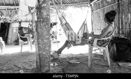 Guna (Kuna) tribal members relax under a large communal hut made of palm fronds, watching as tourists visit their island and shop for Molas Stock Photo