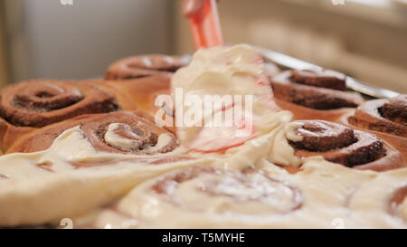 woman or man hand spreads a white cream on freshly baked cinnamon buns with spices with a special spatula in slow motion 4K video. Stock Photo