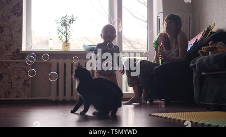 a young beautiful woman blowing soap bubbles inside the room with big window on background and a little baby boy and a black cat are catching this Stock Photo