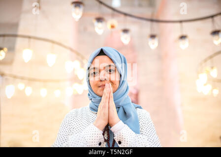 Muslim woman in headscarf and hijab prays while clasping her hands with mosque on background.Religion praying concept. Stock Photo