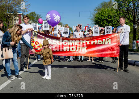 Russia, Nakhodka, 05/09/2017. Nice little girl in military uniform stays in front of big red banner with inscription 'Immortal Regiment'. Annual event Stock Photo