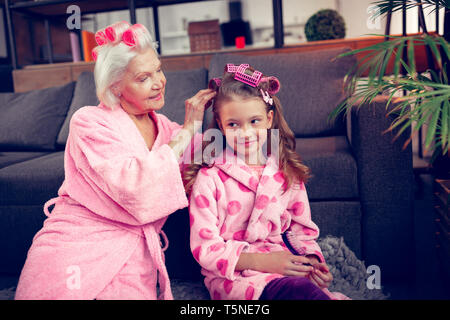 Caring granny putting hair curlers on hair of her cute little girl Stock Photo