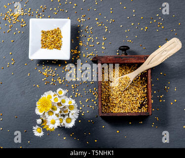 photo composed of a square bowl of white ceramic, a wooden box full of pollen grains of bees and a bunch of daisies, on a slate board, top view Stock Photo