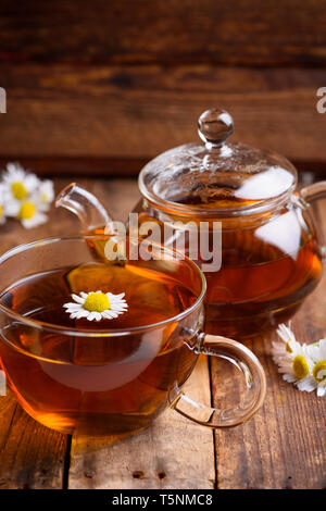 Glass of chamomile tea with full hot teapot and fresh chamomile flowers on vintage old wooden table background Stock Photo