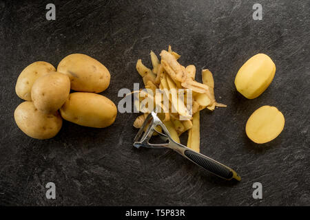 Peeler with peeled potatoes and their skins on textured black background. Photo from above Stock Photo
