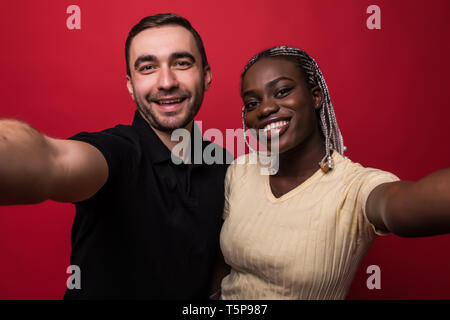 Young interracial couple, caucaisan man and african woman taking selfie on mobile phone on red background Stock Photo