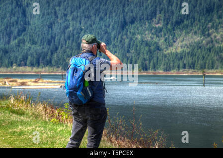 Senior male with backpack looking through binoculars at an Osprey nest.  Pitt Meadows, B. C., Canada Stock Photo