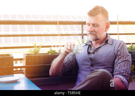 man in his 50s sitting on sunny balcony and drinking coffee Stock Photo