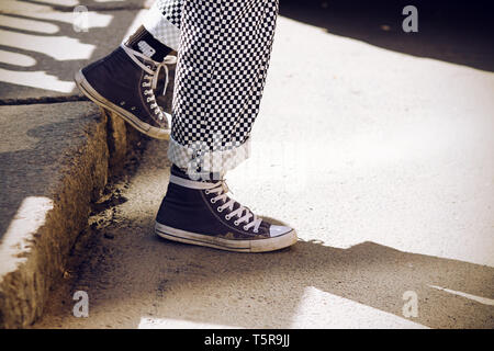 Legs dressed in fashionable retro plaid pants, rolled up at the bottom, and in blue sneakers descend from the step, illuminated by the sunlight Stock Photo