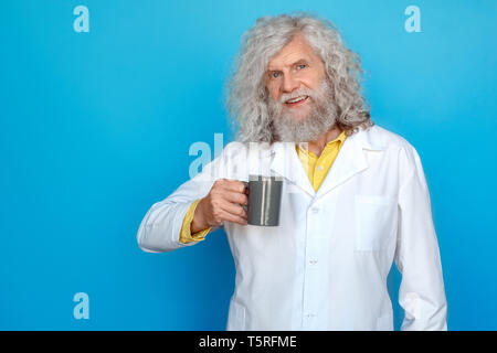 Old long-haired man wearing doctor's gown studio isolated on blue wall standing holding mug of hot coffee looking camera smiling cheerful Stock Photo