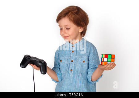 Little boy studio standing isolated on grey wall looking at game controller holding rubik's cube choosing playing console smiling happy Stock Photo