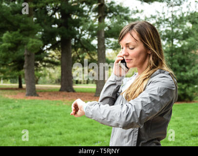 Young woman on cell, mobile phone looking at watch in city park on overcast day, smiling, with green grass, trees background Stock Photo