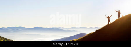Small dark silhouettes of tourist travelers on steep mountain slope at sunrise on copy space background of valley covered with white puffy clouds and  Stock Photo