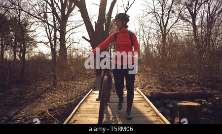 Athlete bicyclist Caucasian woman in sportswear and helmet crosses on foot, leads a mountain bike in her hands across a rural narrow wooden bridge Stock Photo