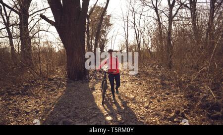 Athlete bicyclist Caucasian woman in sportswear and helmet crosses on foot, leads a mountain bike in her hands across a rural narrow wooden bridge Stock Photo