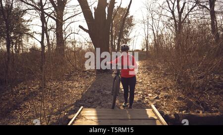 Athlete bicyclist Caucasian woman in sportswear and helmet crosses on foot, leads a mountain bike in her hands across a rural narrow wooden bridge Stock Photo