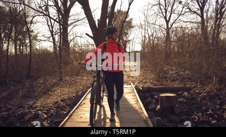 Athlete bicyclist Caucasian woman in sportswear and helmet crosses on foot, leads a mountain bike in her hands across a rural narrow wooden bridge Stock Photo