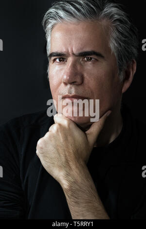 Portrait of a mature man, 56 years old with grey hair, confident, thinking with his hand on chin over black background. Stock Photo
