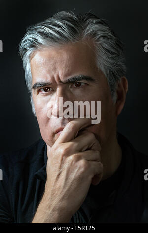 Portrait of a mature man, 56 years old with grey hair, pensive, thinking with his hand on chin over black background, rembrandt lighting. Stock Photo