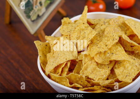 Yellow nacho in white bowl on the wooden table with tomato next to it. Stock Photo