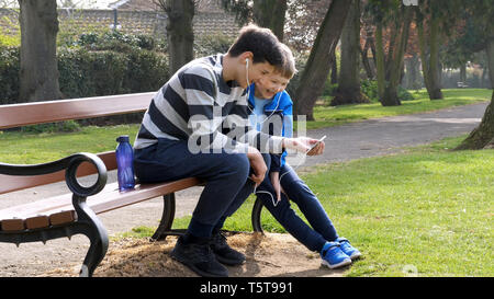 teen boys with smart phone listening or talking in british park on bench. teenager and social media concept. Stock Photo