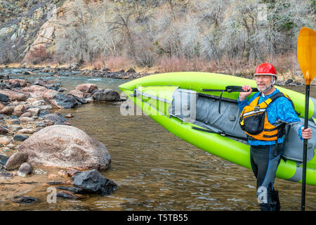 happy senior paddler carrying inflatable whitewater kayak on a shore of mountain river in early spring - Poudre River above Fort Collins, Colorado Stock Photo