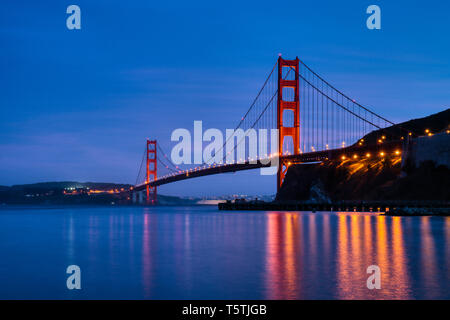 IconicGolden Gate Bridge at blue hour night photograph with twinkling lights and peaceful serene reflection of architecture moody still water Bay Area Stock Photo