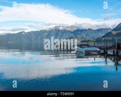 Jet boat at wharf, clouds and blue sky reflected in calm water, distant hills, Lake Hauroko, Fiordland National Park, Southland, New Zealand Stock Photo