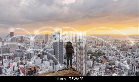 Smart city, wireless network and internet system in the city. Businessman standing on mountain peak with cityscape in sunset Stock Photo
