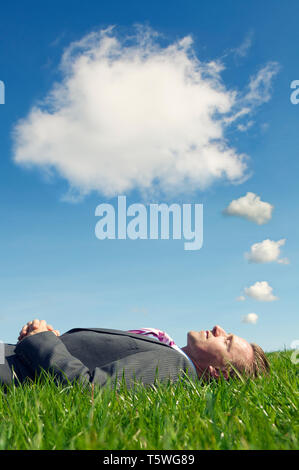 Businessman lying down outdoors in bright green grass dreaming under thought bubble clouds in blue skies Stock Photo