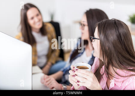 Three young females drinking coffee and chatting in front of desktop computer at the desk. Stock Photo