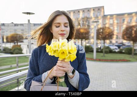 Portrait of young woman with bouquet of yellow spring flowers daffodils. Beautiful girl in city enjoys flowers, eyes closed Stock Photo