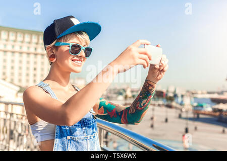 Young alternative girl wearing cap and sunglasses standing on the city street taking photo on smartphone smiling cheerful beautiful view Stock Photo